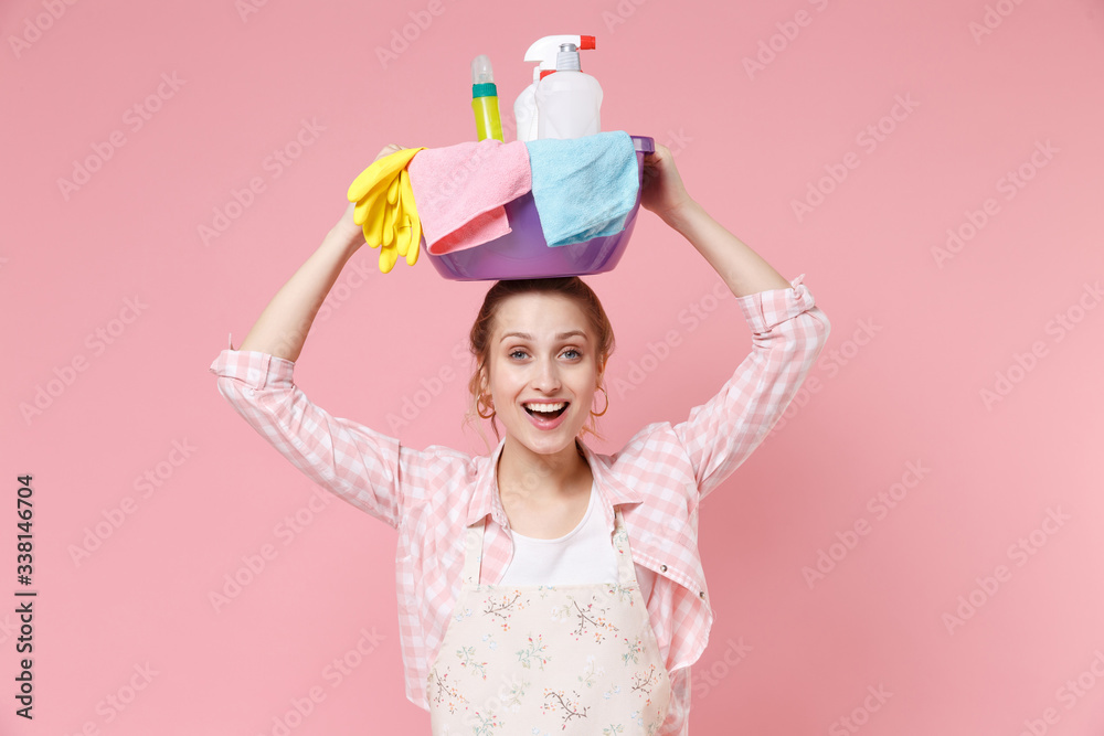Cheerful young woman housewife in apron doing housework isolated on pastel pink wall background studio portrait. Housekeeping concept. Holding basin with detergent bottles washing cleansers on head. Cheerful young woman housewife in apron doing housework isolated on pastel pink wall background studio portrait. Housekeeping concept. Holding basin with detergent bottles washing cleansers on head.