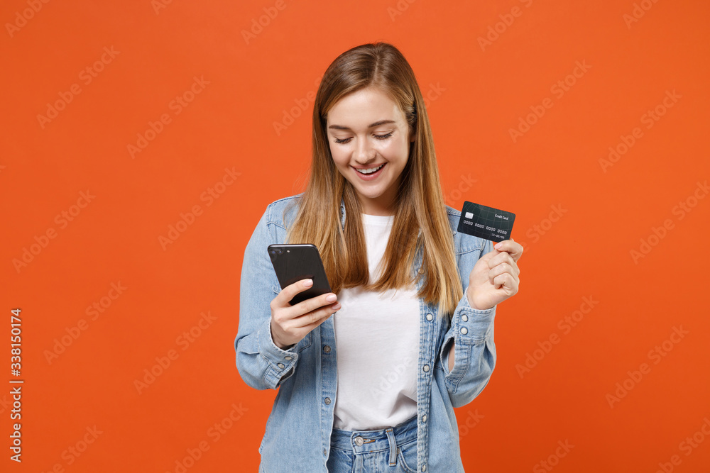 Cheerful pretty young woman girl in casual denim clothes posing isolated on orange wall background studio portrait. People lifestyle concept. Mock up copy space. Using mobile phone, credit bank card.