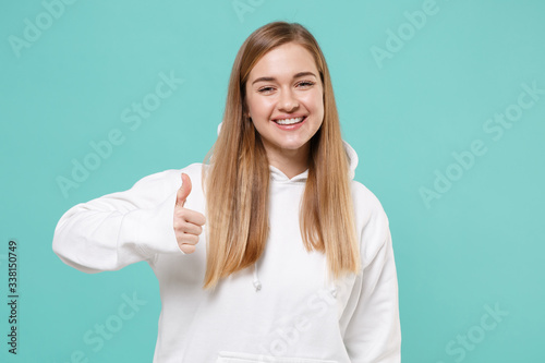 Smiling young woman girl in casual white hoodie posing isolated on blue turquoise wall background studio portrait. People sincere emotions lifestyle concept. Mock up copy space. Showing thumb up.