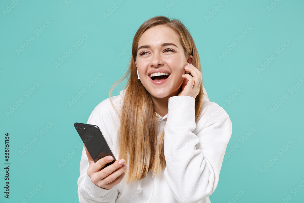 Cheerful young woman in casual white hoodie posing isolated on blue turquoise background studio portrait. People lifestyle concept. Mock up copy space. Listen music with air pods using mobile phone. Cheerful young woman in casual white hoodie posing isolated on blue turquoise background studio portrait. People lifestyle concept. Mock up copy space. Listen music with air pods using mobile phone.