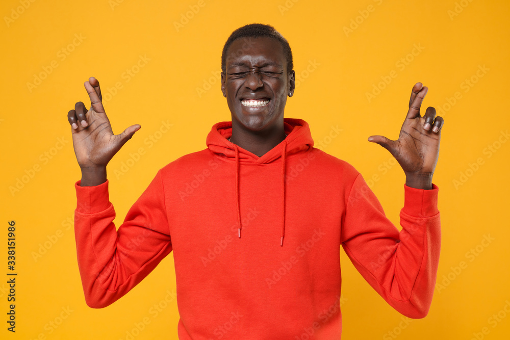 African american man guy in red streetwear hoodie isolated on yellow background. People lifestyle concept. Mock up copy space. Wait for special moment keeping fingers crossed eyes closed, making wish. African american man guy in red streetwear hoodie isolated on yellow background. People lifestyle concept. Mock up copy space. Wait for special moment keeping fingers crossed eyes closed, making wish.