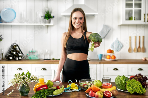 A sporty girl holds .broccoli and stands in the kitchen near a table with fruits and other useful foods. Healthy eating concept