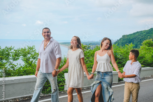 Cheerful, happy family, father, mother, son and daughter enjoying walk play outdoor near sea. Family in white clothes barefoot on walkway. Green forest background. Batumi, Georgia.