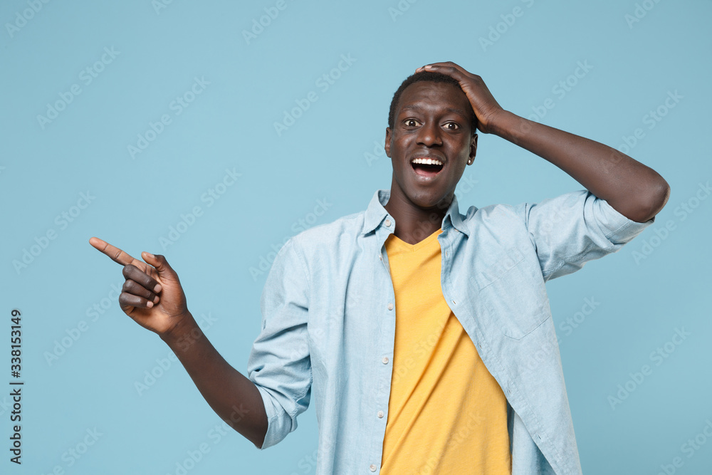 Amazed young african american man guy in casual shirt, yellow t-shirt posing isolated on blue background. People lifestyle concept. Mock up copy space. Pointing index finger aside, put hand on head. Amazed young african american man guy in casual shirt, yellow t-shirt posing isolated on blue background. People lifestyle concept. Mock up copy space. Pointing index finger aside, put hand on head.