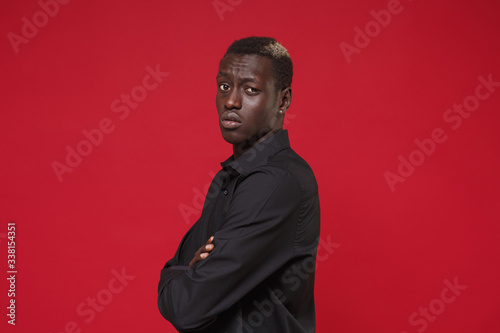 Side view of concerned young african american man in classic black shirt posing isolated on red wall background. People lifestyle concept. Mock up copy space. Holding hands crossed, looking camera.