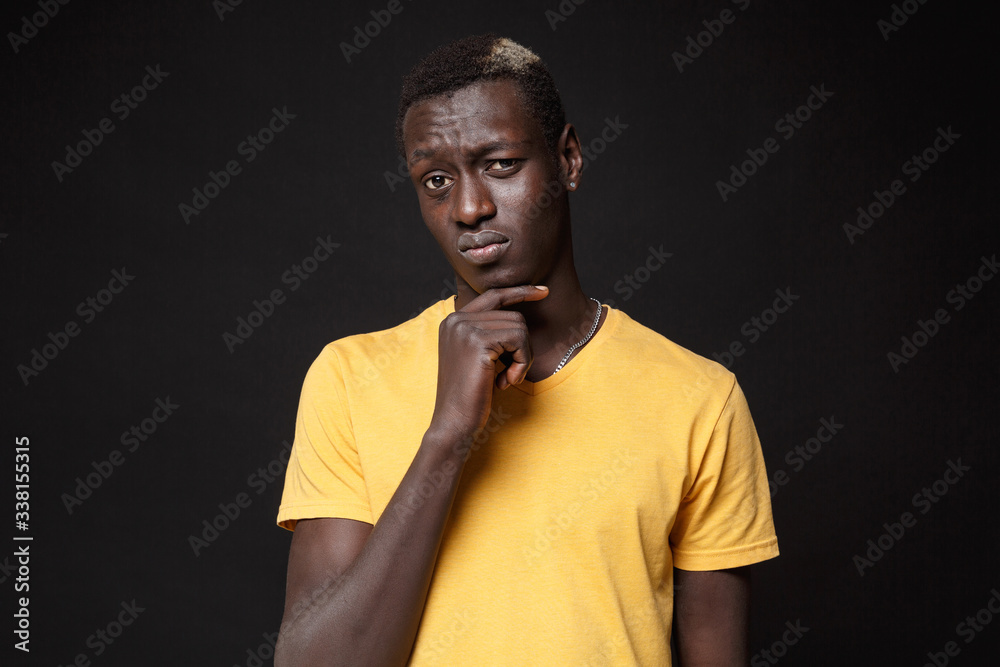 Puzzled young african american man guy in yellow t-shirt posing isolated on black background studio portrait. People sincere emotions, lifestyle concept. Mock up copy space. Put hand prop up on chin.