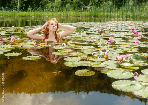 beautiful young sexy red-haired woman mermaid stretches sensually and seductively in the water  lake with pink water lilies and strokes her hair