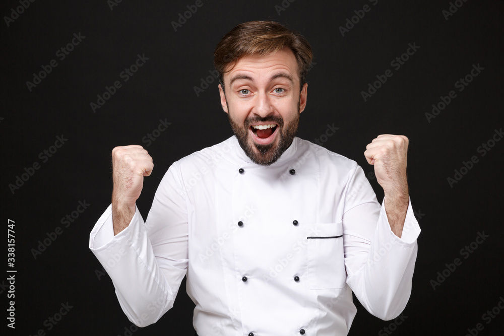 Surprised young bearded male chef cook or baker man in white uniform shirt posing isolated on black background studio portrait. Cooking food concept. Mock up copy space. Clenching fists like winner. Surprised young bearded male chef cook or baker man in white uniform shirt posing isolated on black background studio portrait. Cooking food concept. Mock up copy space. Clenching fists like winner.