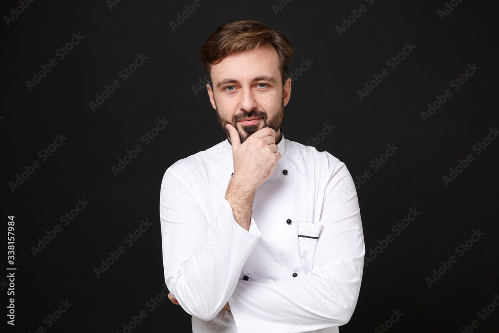 Handsome young bearded male chef cook or baker man in white uniform shirt posing isolated on black wall background studio portrait. Cooking food concept. Mock up copy space. Put hand prop up on chin. Handsome young bearded male chef cook or baker man in white uniform shirt posing isolated on black wall background studio portrait. Cooking food concept. Mock up copy space. Put hand prop up on chin.