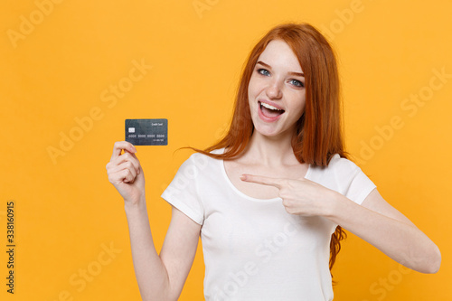 Excited young redhead woman girl in white blank empty t-shirt posing isolated on yellow background in studio. People lifestyle concept. Mock up copy space. Pointing index finger on credit bank card.