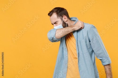 Young man in sterile face mask posing isolated on yellow wall background. Epidemic pandemic spreading coronavirus 2019-ncov sars covid-19 flu virus concept. Coughing or sneezing, covering with elbow. photo