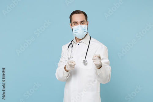 Male doctor man in white medical gown sterile face mask gloves isolated on blue background. Epidemic pandemic coronavirus 2019-ncov sars covid-19 flu virus concept. Pointing index fingers on camera.