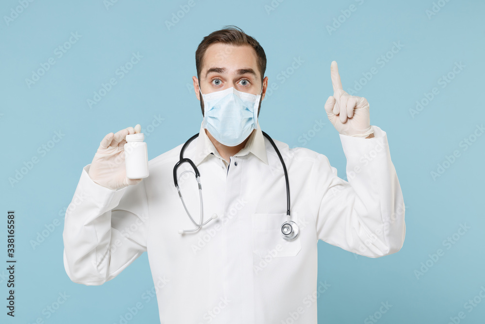 Doctor man in gown face mask gloves isolated on blue background. Epidemic pandemic coronavirus 2019-ncov sars covid-19 flu virus concept. Hold medication tablets pills in bottle point index finger up.