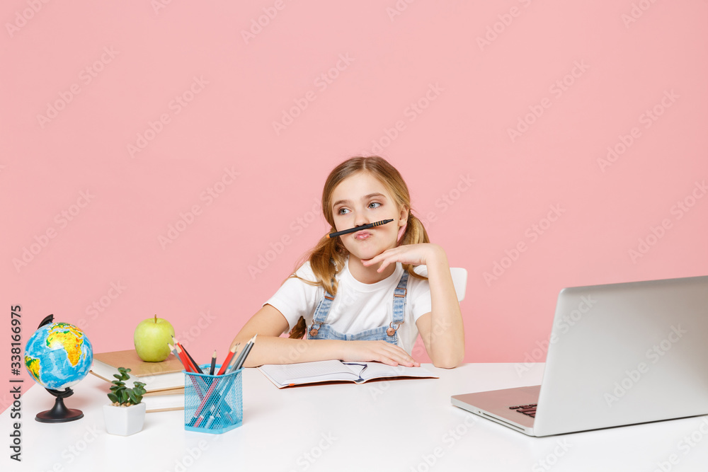 Boring little kid schoolgirl 12-13 years old in white t-shirt sit studying at white desk with pc laptop isolated on pastel pink background. School distance education at home during quarantine concept. Boring little kid schoolgirl 12-13 years old in white t-shirt sit studying at white desk with pc laptop isolated on pastel pink background. School distance education at home during quarantine concept.