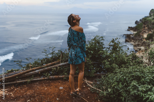 A beautiful girl stands on a cliff overlooking the waves of the blue ocean. Tropical paradise and exotic nature on the island of Bali. Model with different emotions of happiness and joy. photo