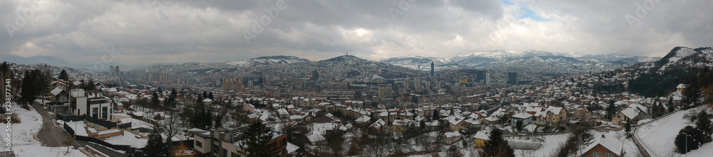 Winter drone panorama of Sarajevo, capital of Bosnia and Herzegovina, taken on a snowy winter day with clouds. Snow is seen on the houses and the mountains in the back.