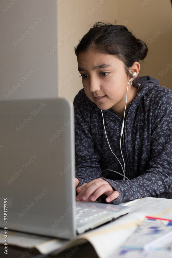 Young girl sitting at the dining table with laptop at home schooling, online virtual classroom video conference, distant education. Active participation at the lesson with earphones, doing homework. Young girl sitting at the dining table with laptop at home schooling, online virtual classroom video conference, distant education. Active participation at the lesson with earphones, doing homework.