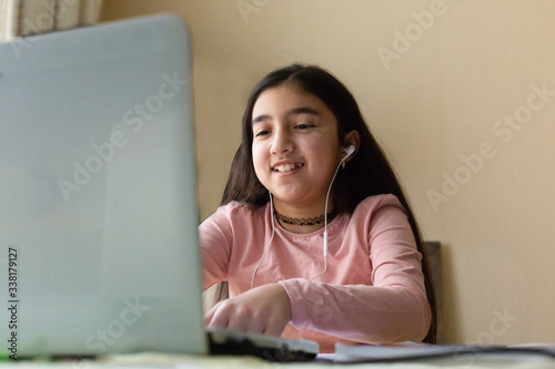 Young girl sitting at the dining table with laptop at home schooling, online virtual classroom video conference, distant education. Active participation at the lesson with earphones, doing homework.