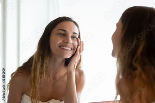 Happy young woman wrapped in towel after showering looking in mirror, touching under eyes area, feeling satisfied with antiwrinkle product effect. Smiling millennial lady doing daily skincare routine. photo