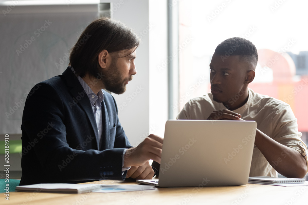 Serious multiracial male colleagues sit at office desk brainstorm talk discuss financial ideas working on laptop together, diverse coworkers cooperate at briefing using computer, collaboration concept Serious multiracial male colleagues sit at office desk brainstorm talk discuss financial ideas working on laptop together, diverse coworkers cooperate at briefing using computer, collaboration concept