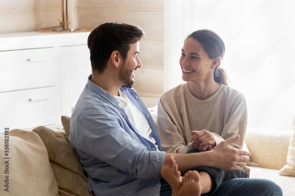 Obraz premium Happy young couple resting on comfortable sofa, chatting at home. Smiling mixed race girl putting legs on boyfriend, relaxing on couch in living room, enjoying sincere conversation, dating concept.