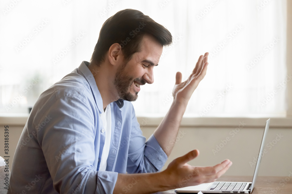 Side view excited young man looking at laptop screen, reading email with unbelievable good news. Cheerful bearded guy amazed by good job results, online lottery win, sitting at table at home. Side view excited young man looking at laptop screen, reading email with unbelievable good news. Cheerful bearded guy amazed by good job results, online lottery win, sitting at table at home.