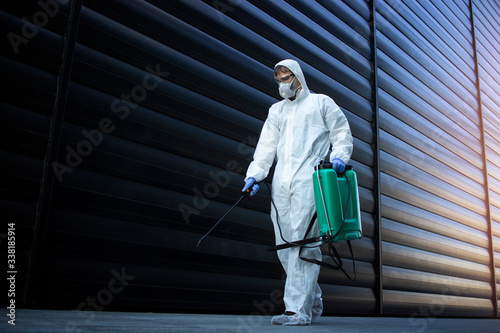 Shot of male person in white chemical protection suit doing disinfection of public areas to stop spreading highly contagious corona virus. Stop coronavirus or COVID-19.