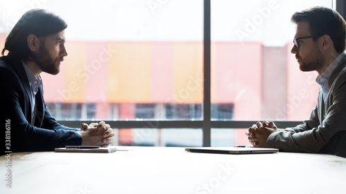 Confident Caucasian businessmen sit opposite at office desk debate in boardroom, serious male business opponents or competitors look at each other at briefing, rivalry, confrontation concept