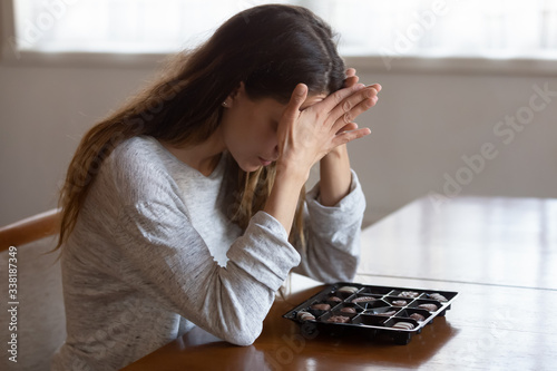 Quadro em tela Depressed young mixed race woman sitting at table with chocolate sweets, feeling desperate alone at home