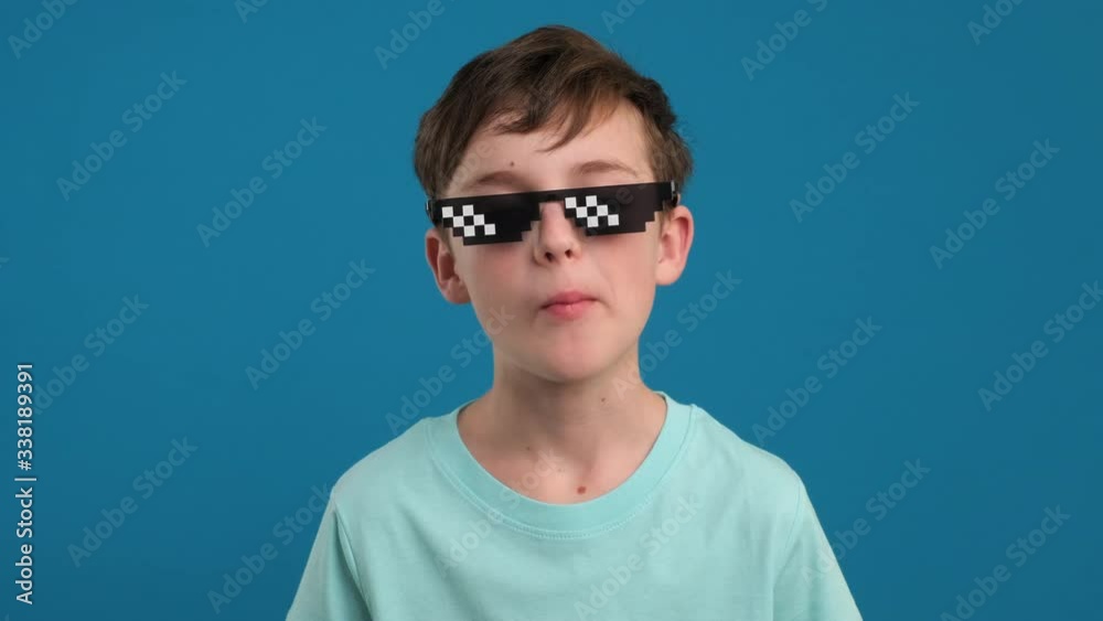 Portrait happy boy chewing gum and learning to inflate bubbles and bursting in pixel sunglasses it joyfully smiling looking at camera slow motion on blue background. Childhood. Emotions people