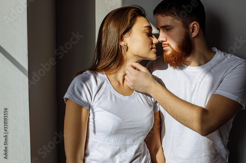 portrait of young and beautiful married couple in love. attractive caucasian couple stand near window, they feel trembling tender feelings for each other, rays of the sun fall on skin, going to kiss