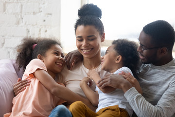 African american happy family with two kids enjoy time together. Cute diverse daughter and son with parents sit on warm comfy couch in living room. Young father and mother have fun with adorable kids