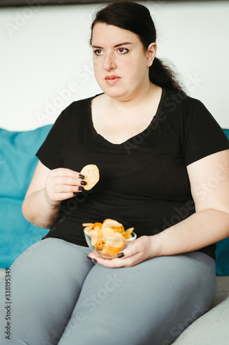 Overeating and gluttony concept. Mindless snacking  home sedentary lifestyle. Obese woman sitting on sofa eating unhealthy food