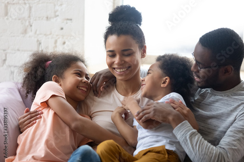 African american happy family with two kids enjoy time together. Cute diverse daughter and son with parents sit on warm comfy couch in living room. Young father and mother have fun with adorable kids photo