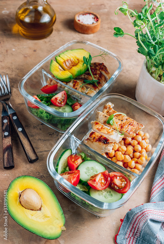 Healthy meal prep containers with chickpeas, chicken, tomatoes, cucumbers and avocados. Healthy lunch in glass containers on beige rustic background. Zero waste concept. Selective focus.