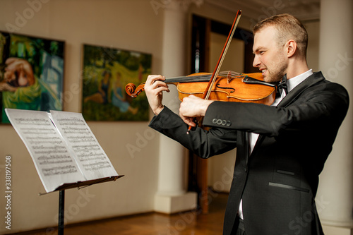 young caucasian man in formal suit play violin, look at notes and gracefully perform music. indoors