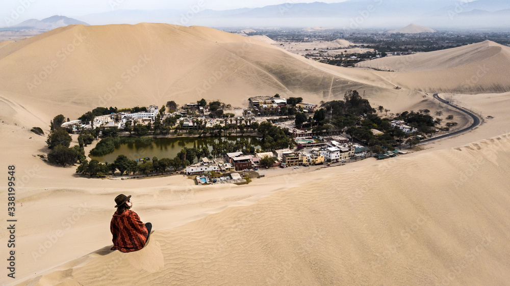 Aerial view on a solo female traveler sitting on the sand dune wearing an Andean styled poncho and brown hat with the Huacachina Oaza in the background. Huacachina, Ica, Peru, South America. 