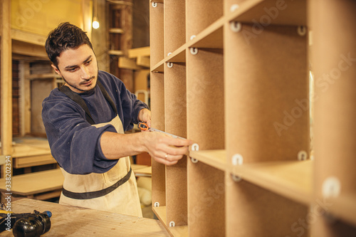 custom-made furniture concept. handicraft carpentry. young caucasian woodworker measuring a hand made wooden shelves using tape-line in factory