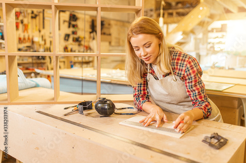 attractive caucasian carpenter lady deals with drawings during work, woman use pencil and ruler before making wooden furniture in workshop