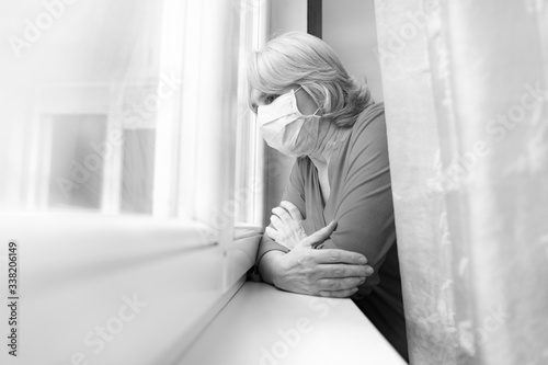 Sad senior woman looking outside her window with mask on face, coronavirus © phoenix021