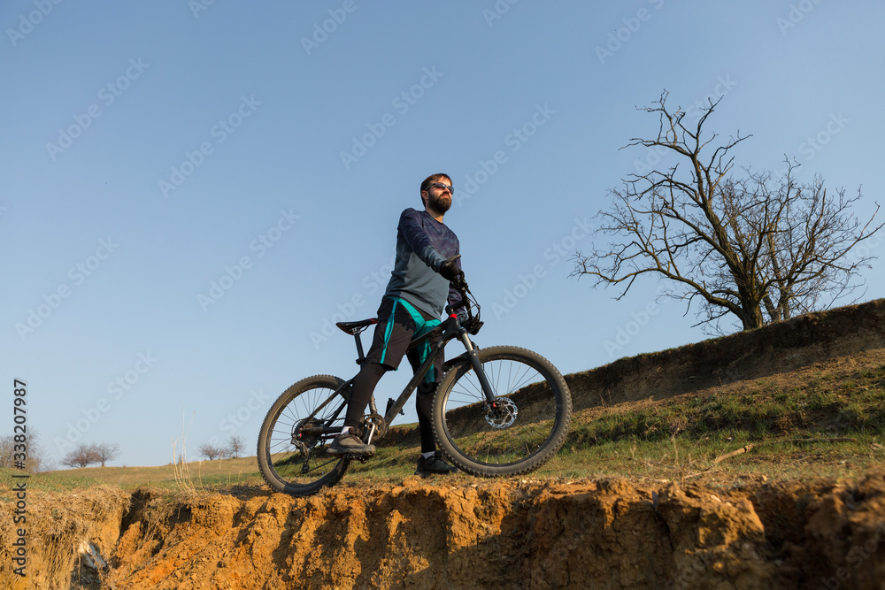 Obraz premium Cyclist in shorts and jersey on a modern carbon hardtail bike with an air suspension fork standing on a cliff against the background of fresh green spring forest