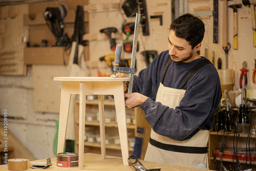 portrait of young caucasian carpenter making wooden chair in the factory, wearing uniform, man keen on handicraft