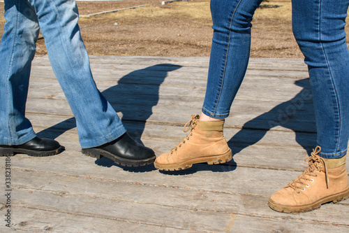 Two people kick each other. Denim pants, boots. Wooden background outdoors. Concept of greetings using leg, social distance during a covid-19 virus pandemic.
