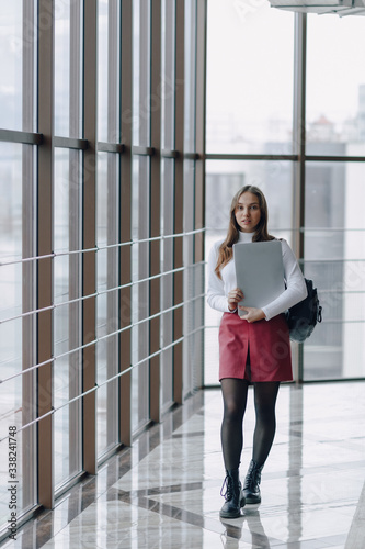attractive girl walks in a bright corridor with laptop and things in airport terminal or office. travel atmosphere or alternative work atmosphere. freelancer student travels to business meeting.