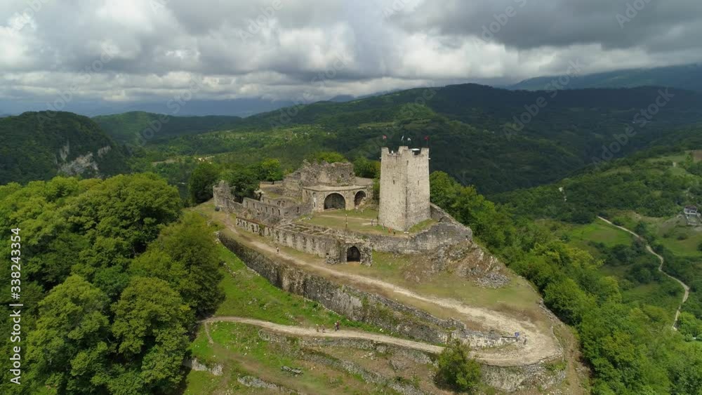 Fortress on sharp mountain ancient stone stronghold Georgia unique landmark on top of mountain. Natural landscape cityscape. Cumulus dramatic clouds. People tourists on tower. Aerial forward flight Fortress on sharp mountain ancient stone stronghold Georgia unique landmark on top of mountain. Natural landscape cityscape. Cumulus dramatic clouds. People tourists on tower. Aerial forward flight