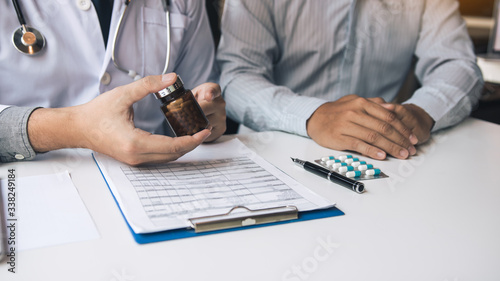 Confident doctor man holding a pill bottle and writing while talking with senior patient and reviewing his medication at office room. photo