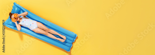 Top view on a little girl in a swimsuit and half glasses of orange resting on an inflatable blue mattress on a yellow background, concept of summer vacation. Banner copy space photo