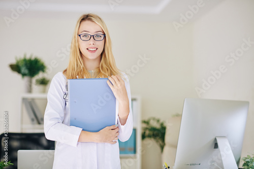 Portrait of beautiful blond young general practitioner in glasses holding fodler and smiling at camera photo
