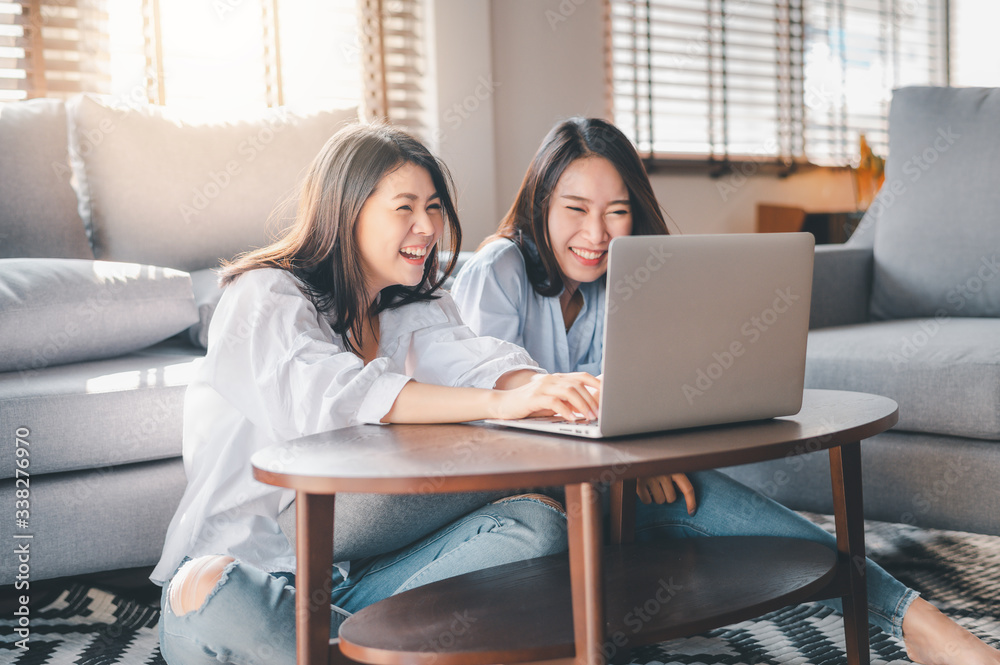 Two happy Asian women best friends in casual wear laughing while working with laptop at home in living room Two happy Asian women best friends in casual wear laughing while working with laptop at home in living room