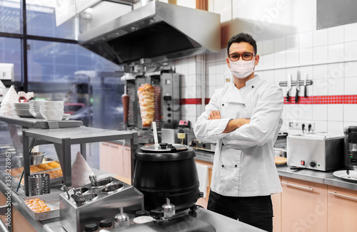 health protection, safety and pandemic concept - male chef cook wearing face protective medical mask over kebab shop kitchen background